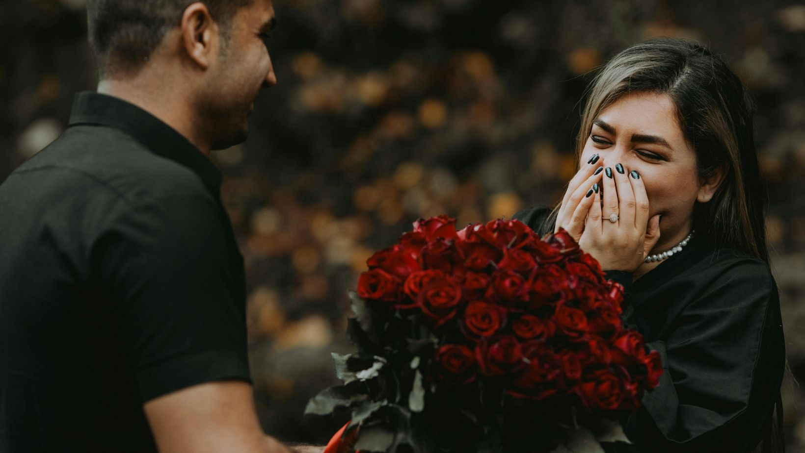 A man in a black shirt surprises a smiling woman with a large bouquet of red roses. She covers her mouth in excitement, reacting with joy. The background is blurred, creating a romantic and emotional atmosphere.